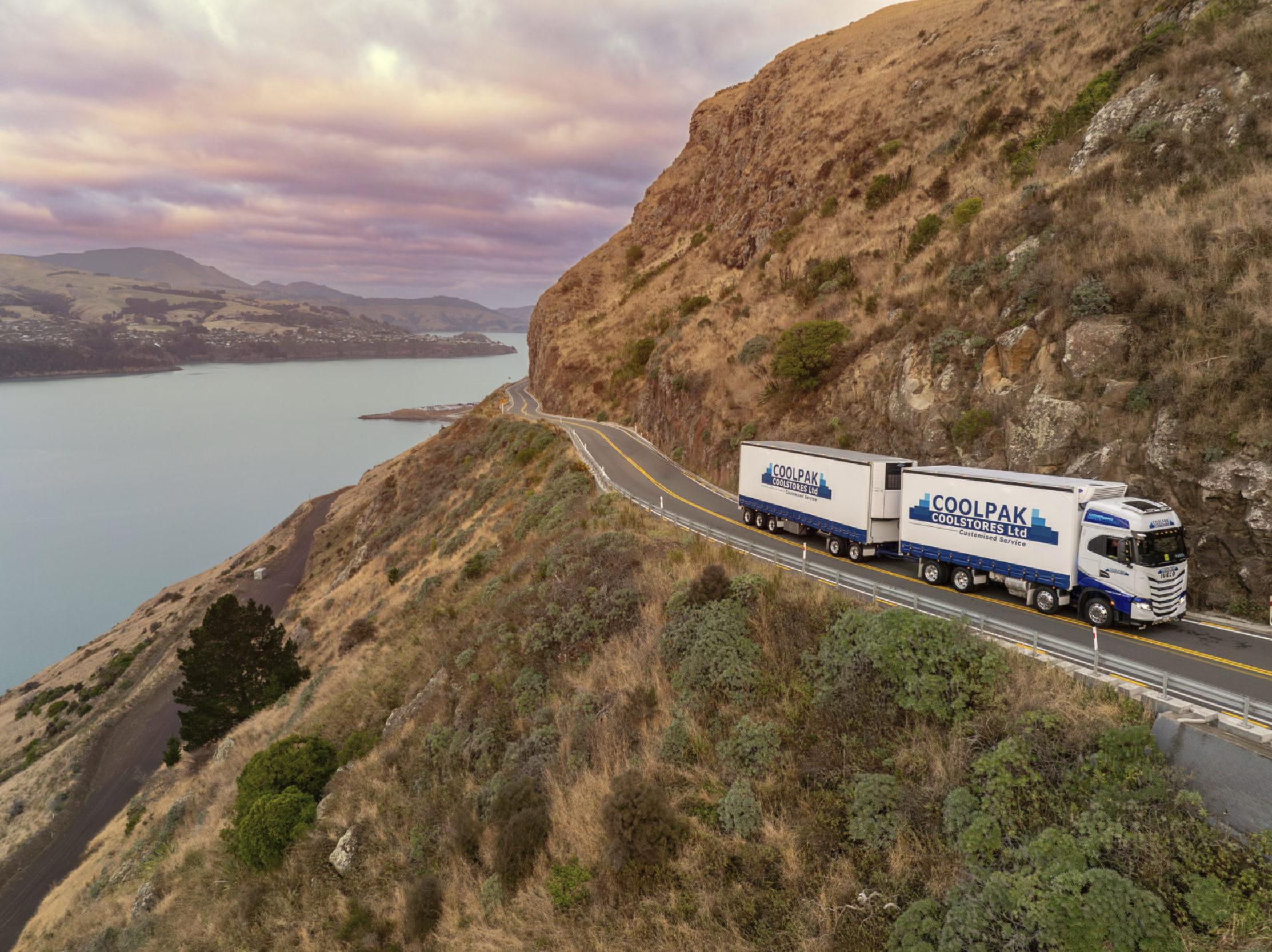A large white and blue semi-truck labelled CoolPak Coolstores Ltd driving along a winding coastal road surrounded by hills and water under a cloudy sky